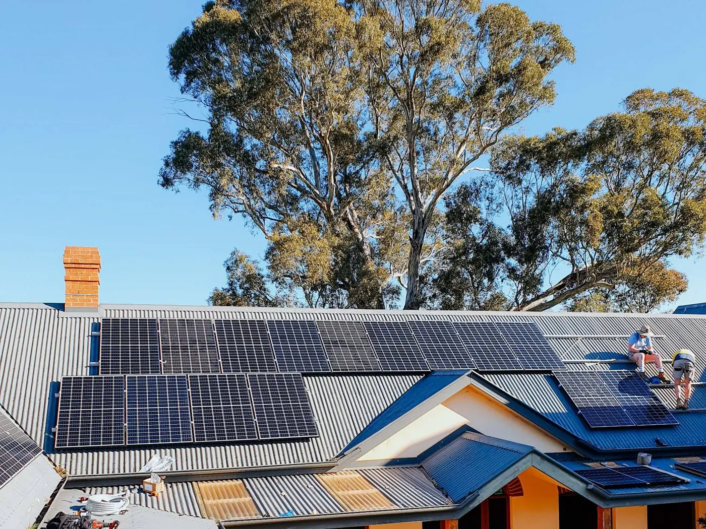 Installers fitting solar panels on the corrugated iron roof of an older home beside tall gum trees