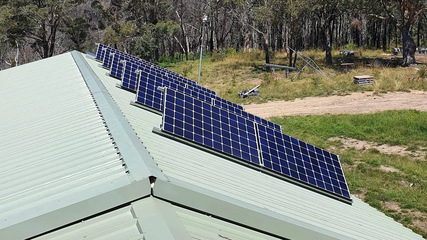 Rows of tilt-framed solar panels on a green corrugated iron roof, with bushland behind